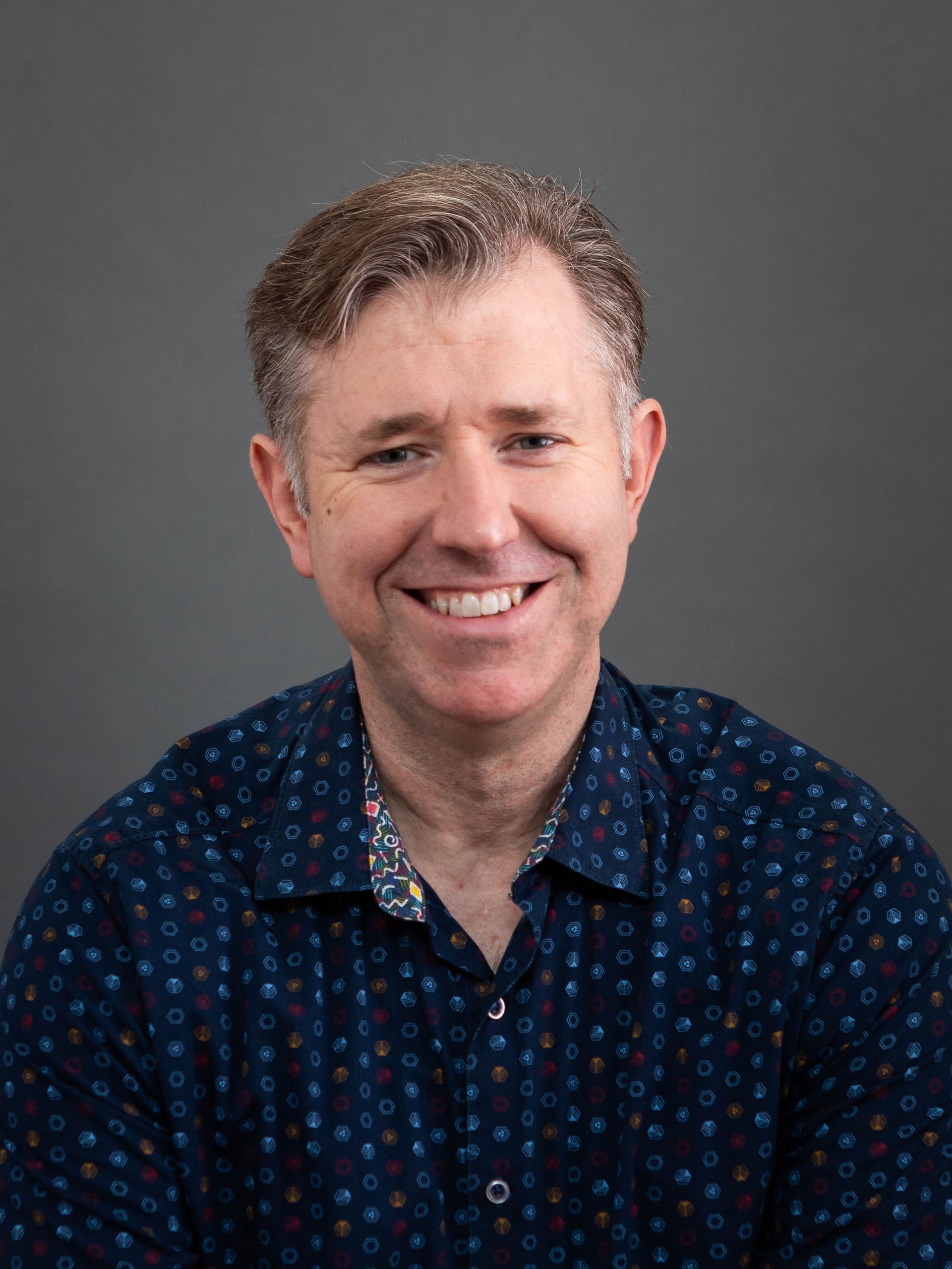 A photo of John McKoll wearing a blue patterned button-down shirt in front of a gray background.